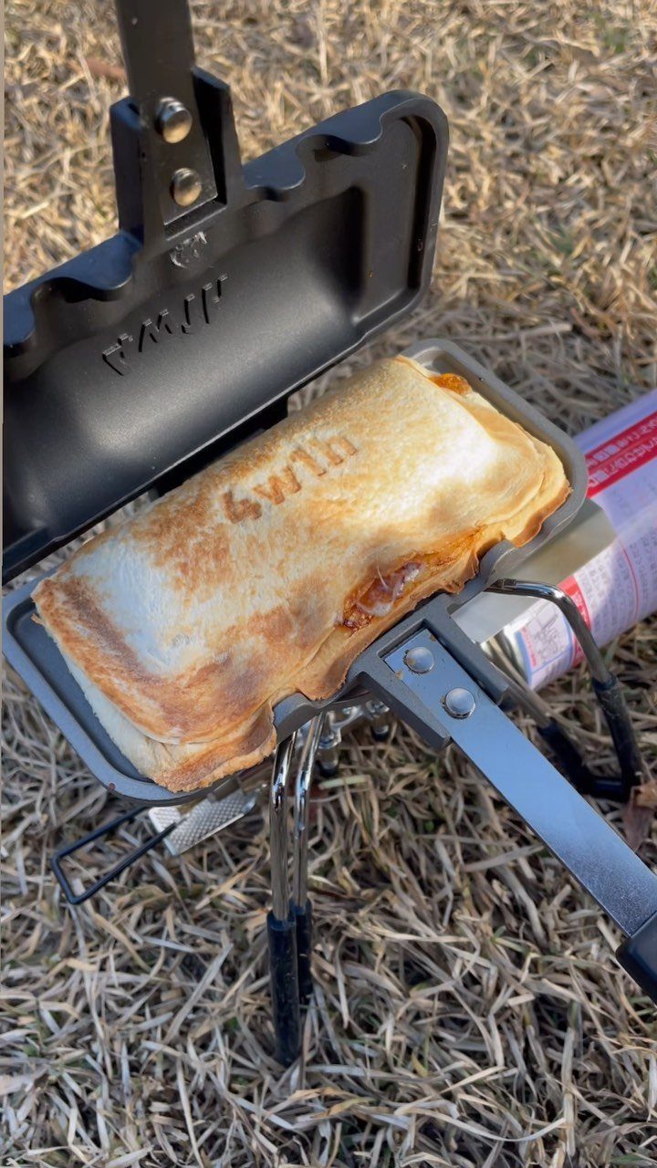 Toasted grilled cheese sandwich inside a black metal campfire press on dry grass.