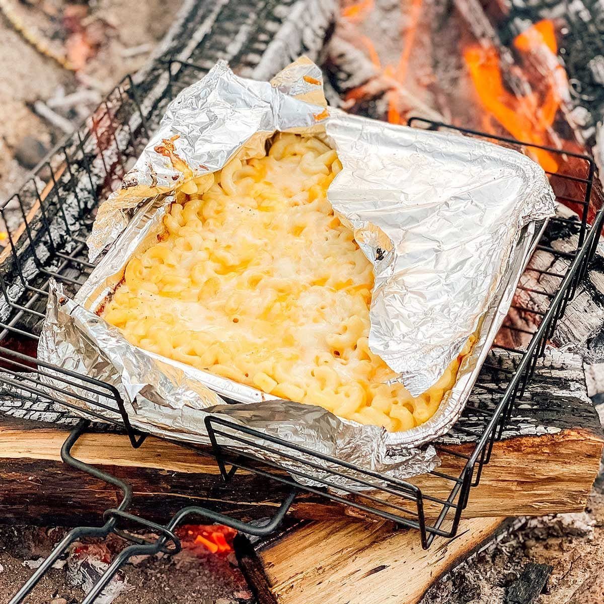 Macaroni and cheese in a foil pan cooking over a campfire grill grate.
