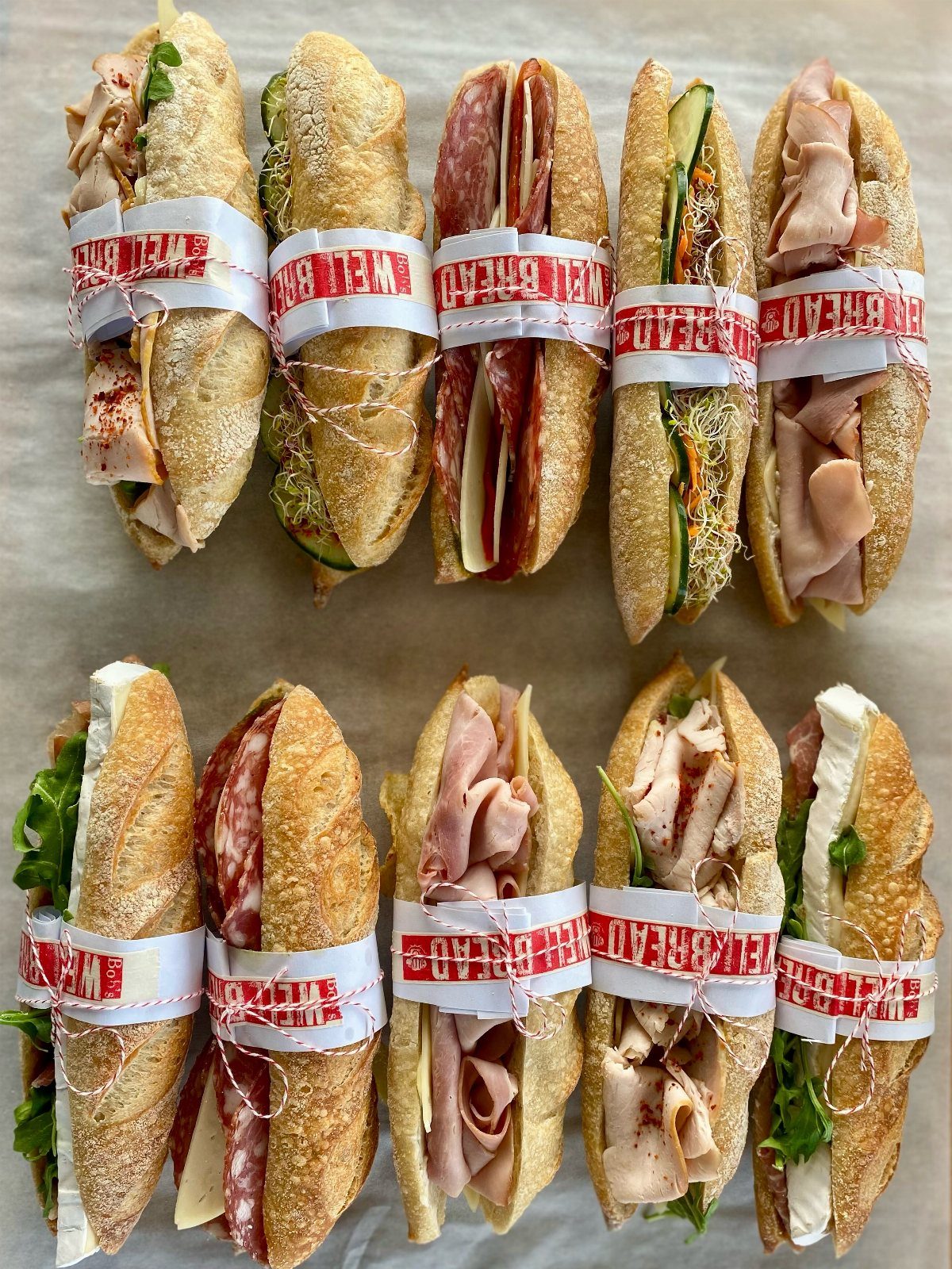 Assorted baguette sandwiches wrapped in red and white paper bands on white paper.