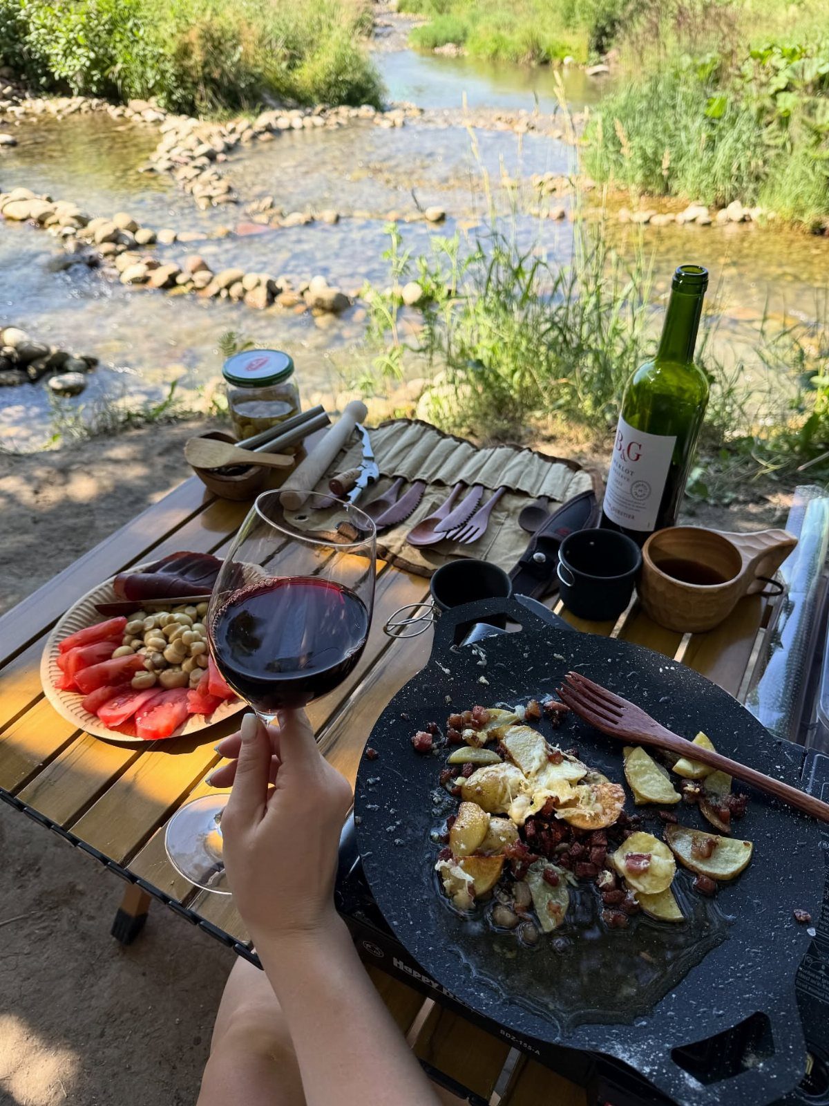 Red wine glass held over a camping table with a potato skillet by a stream