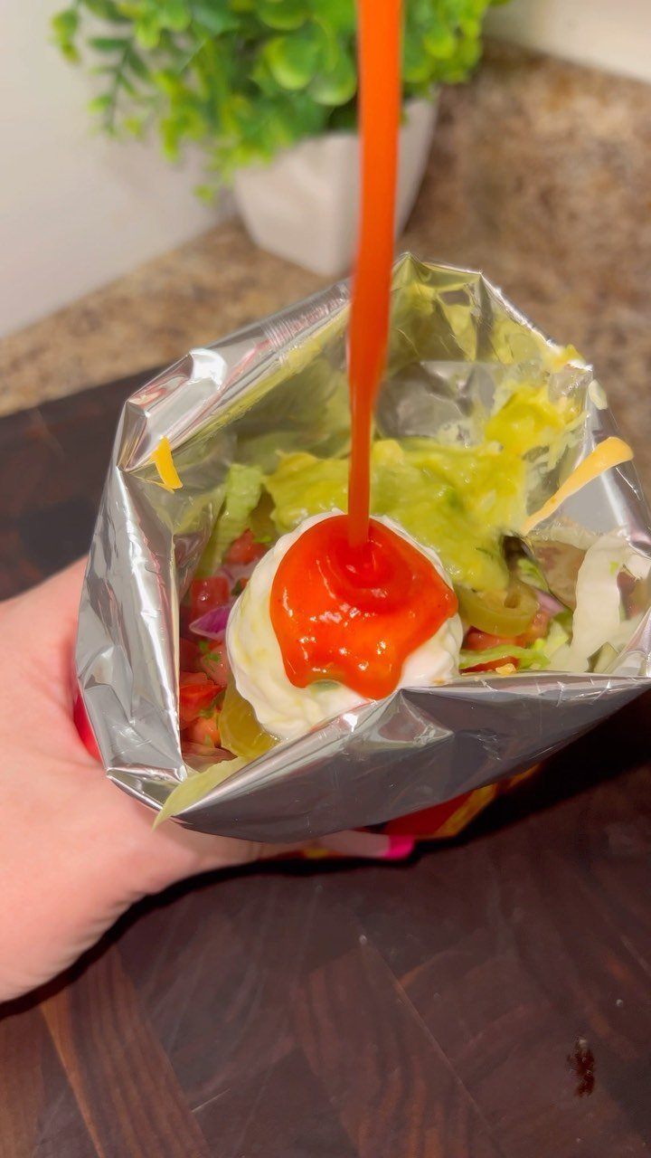 Taco ingredients in a silver foil bag with orange sauce being poured over sour cream