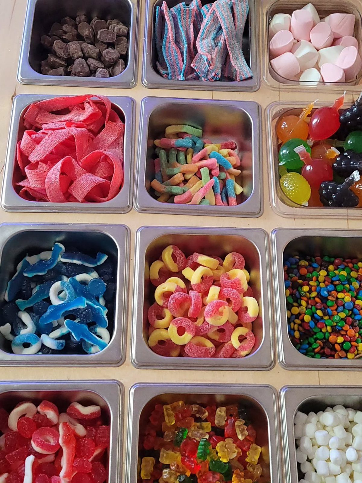 Colorful candies in square stainless steel containers arranged in a grid on wood.