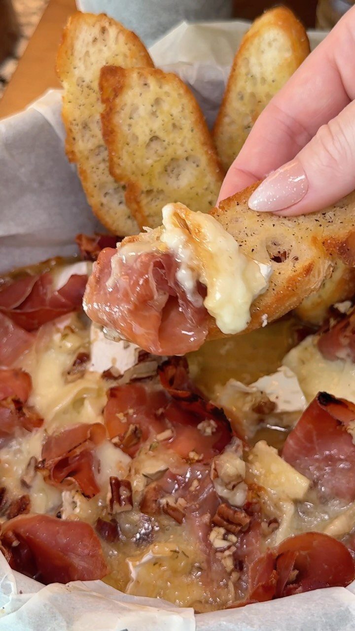 Melted baked brie with prosciutto and pecans being scooped with toasted baguette slices.