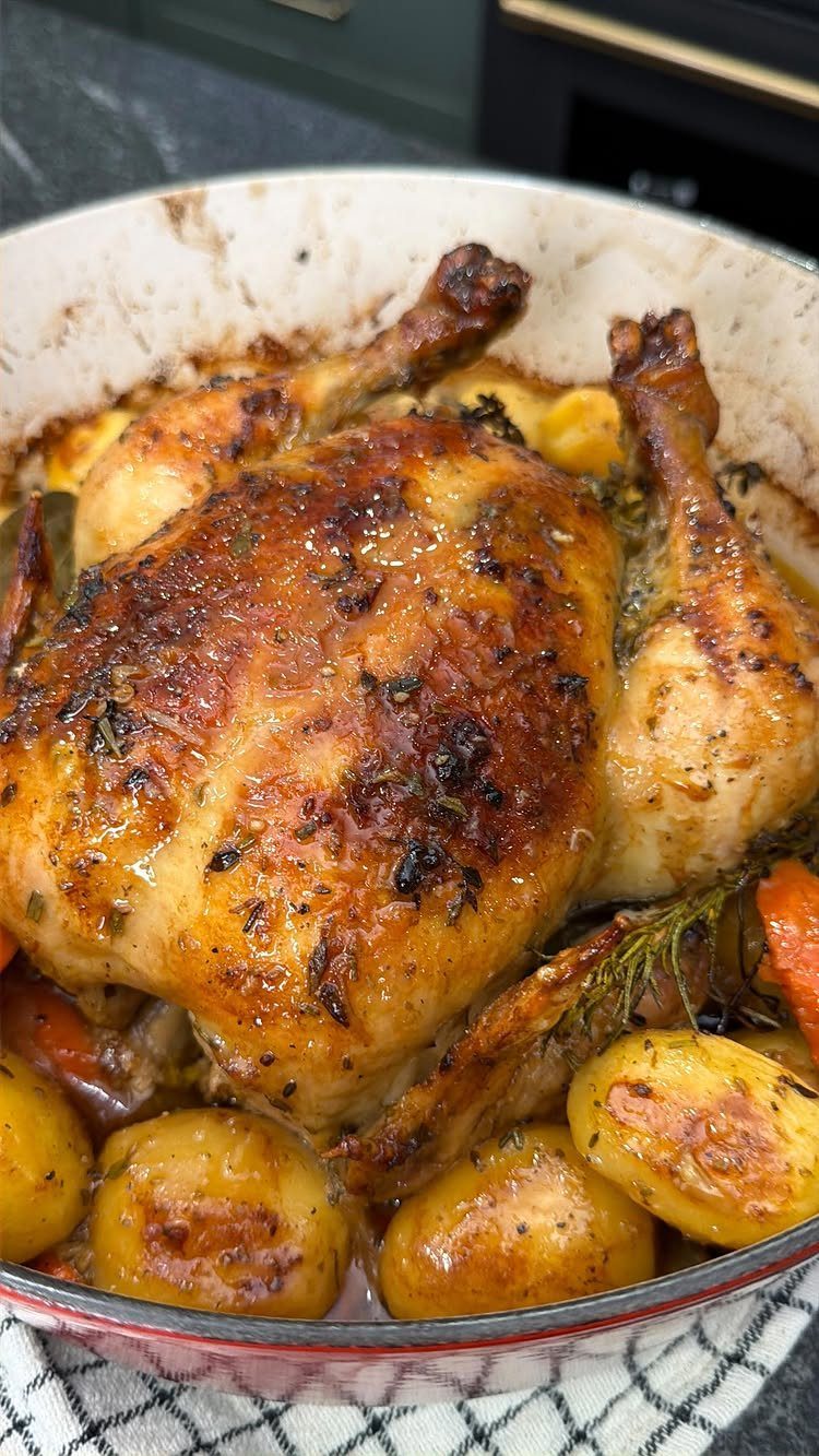 Whole roasted chicken with golden skin and roasted potatoes in a white pan.