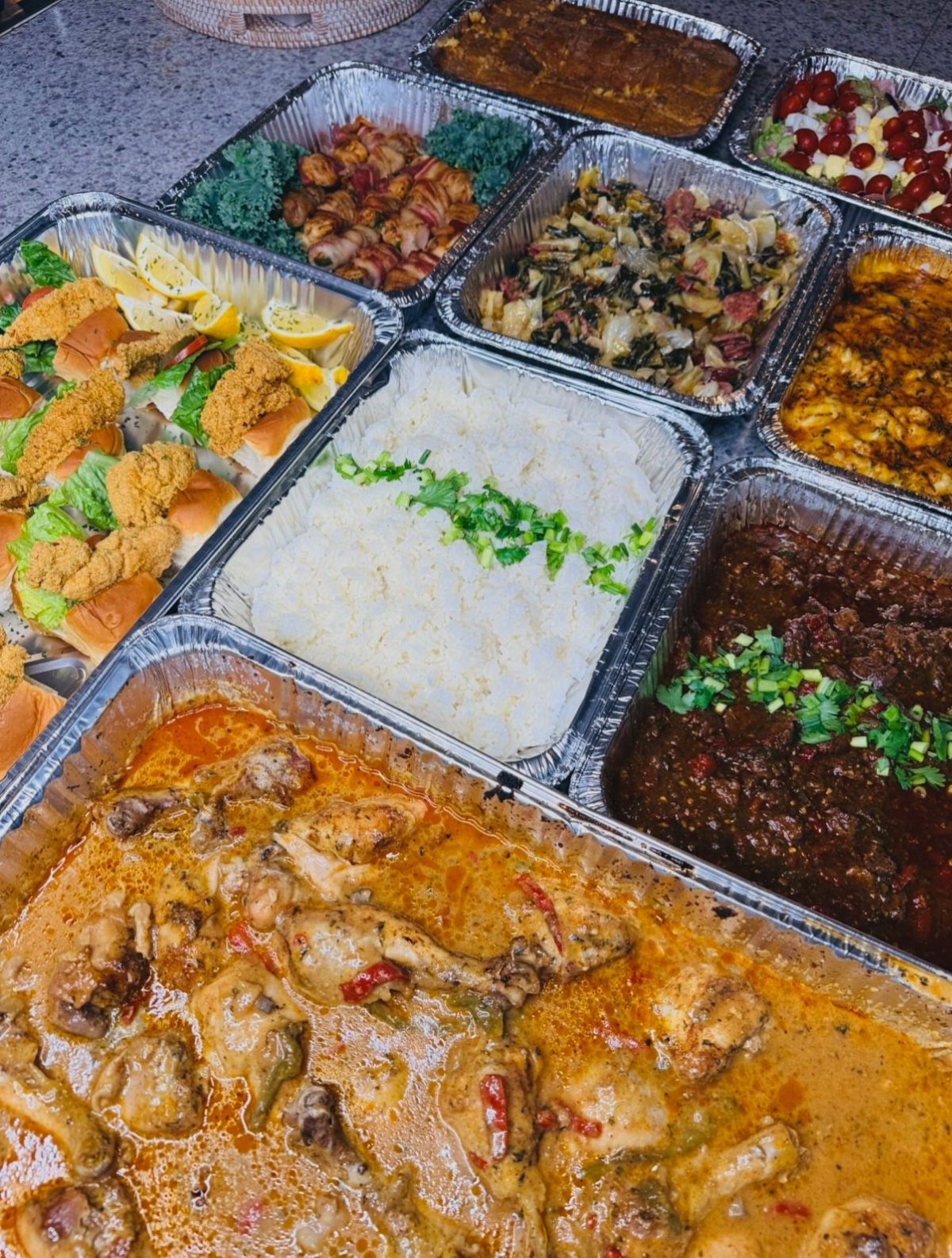 Assorted catered food dishes in aluminum trays arranged on a grey surface.