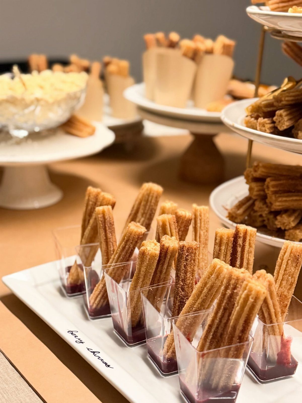 Mini churros in clear cups with red sauce on a white dessert platter.