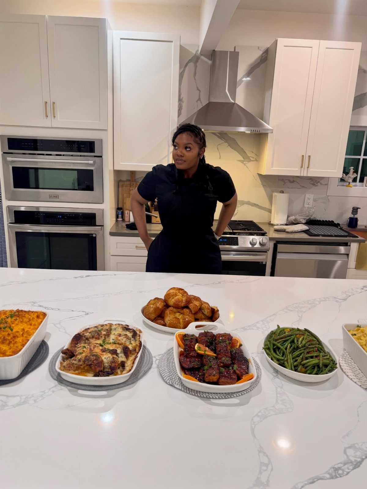 Female chef standing behind a marble counter with various gourmet prepared dinner dishes.