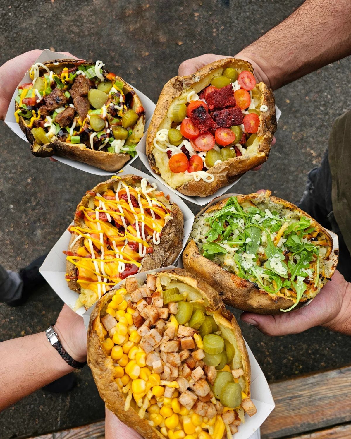 Five loaded baked potatoes with diverse toppings held in white cardboard trays.