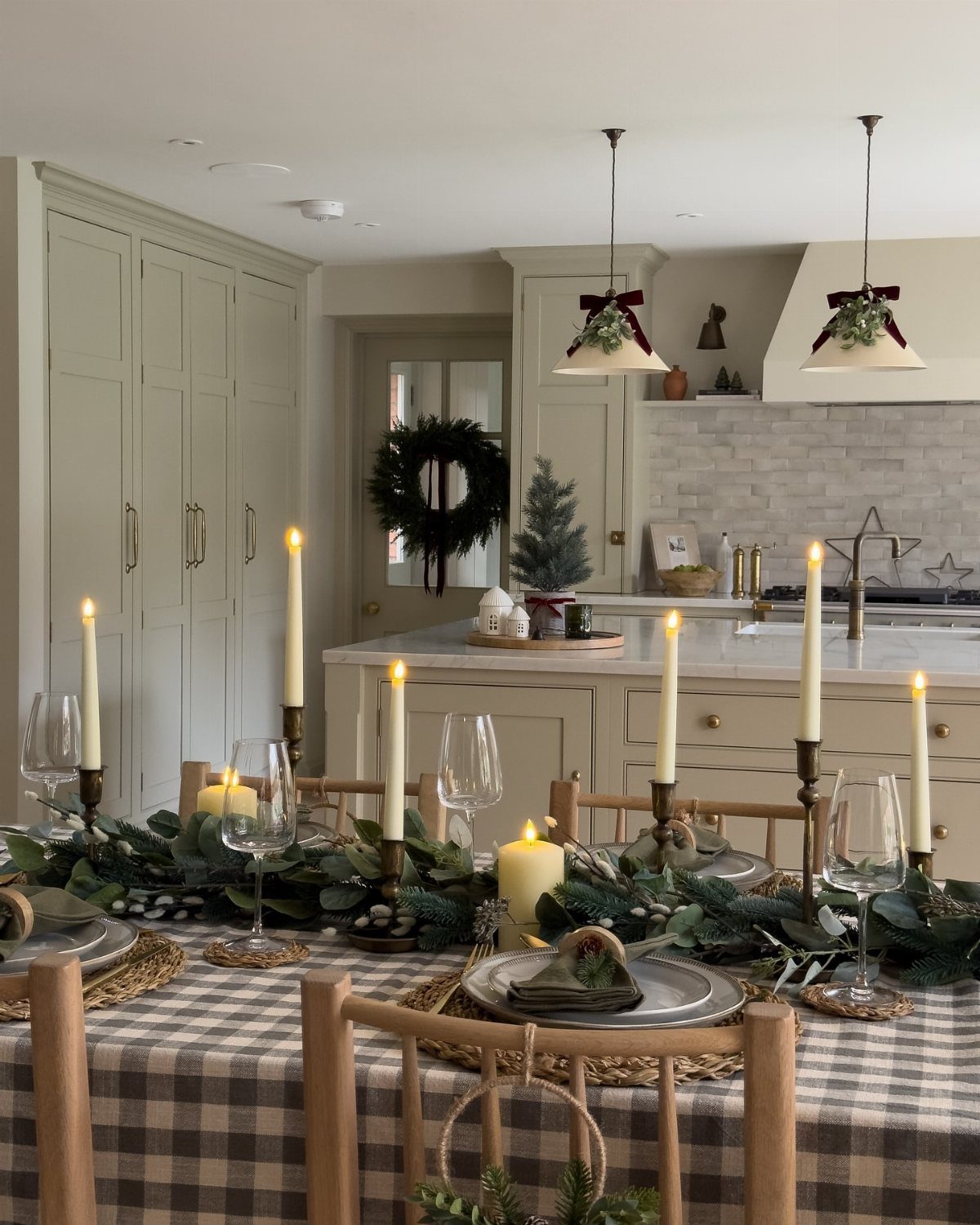 Dining table with checkered cloth, greenery garland, and lit white candles in kitchen.