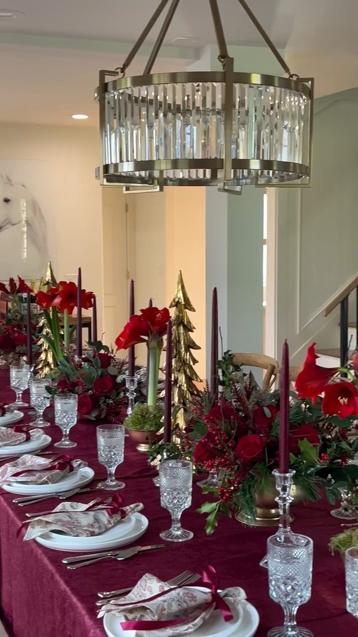 Festive dining table with red tablecloth, red flowers, green foliage, and gold accents.