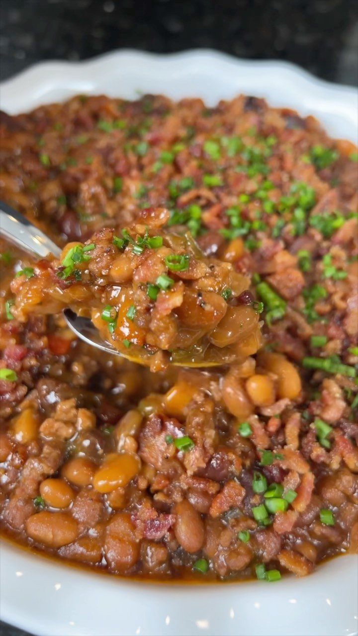 Baked beans with bacon and chives in a white bowl being scooped by spoon