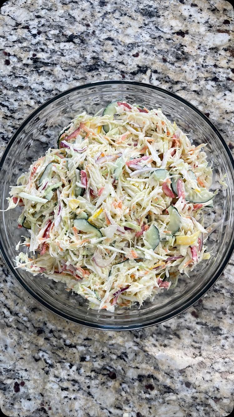 Creamy coleslaw with shredded cabbage and cucumbers in a glass bowl on granite.