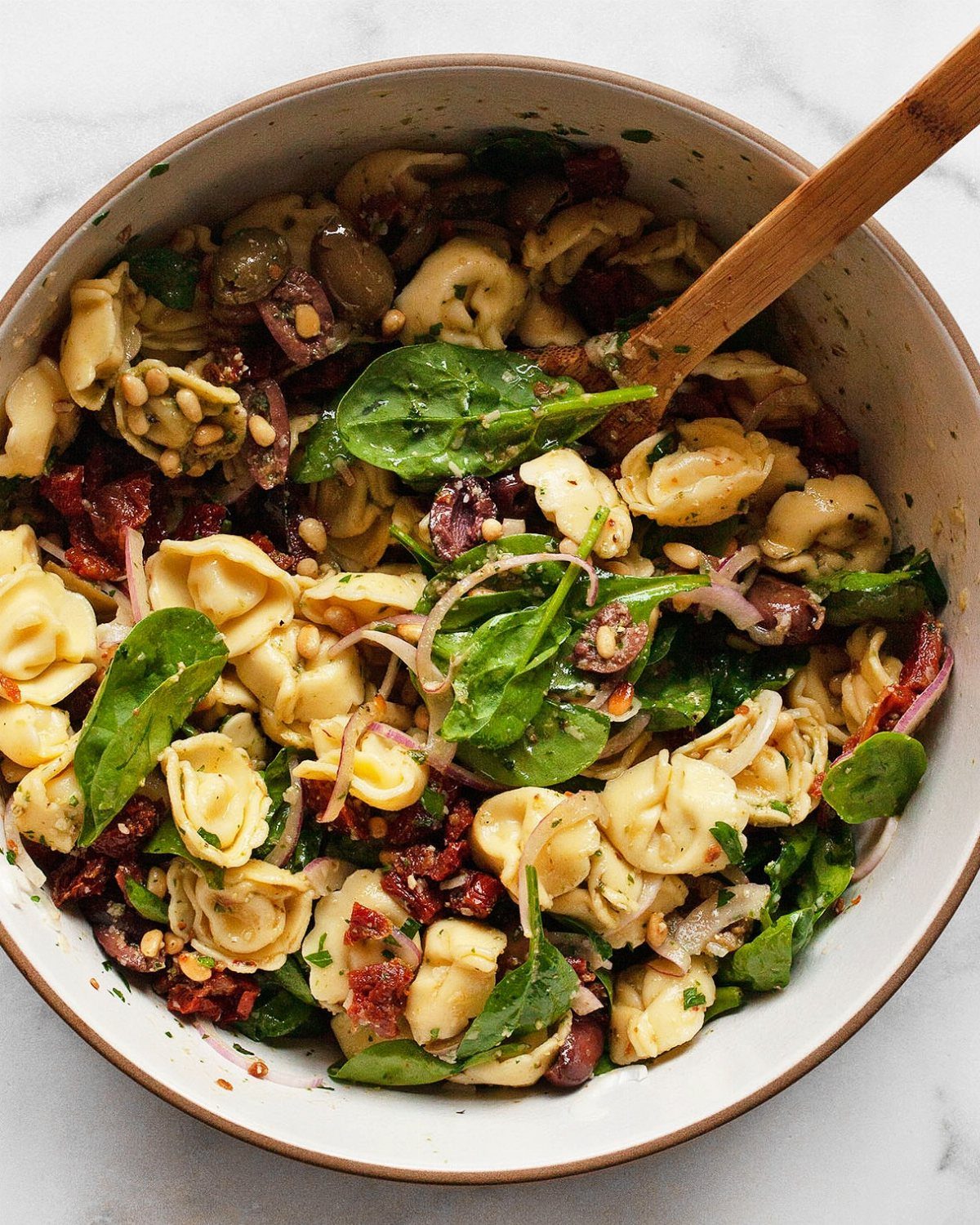 Tortellini pasta salad with spinach, sun-dried tomatoes, and olives in a brown bowl.
