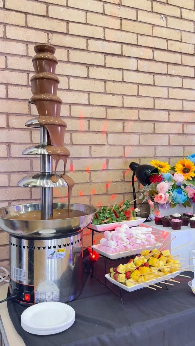 Chocolate fountain with flowing chocolate, fruit platters, and marshmallows on a black table.