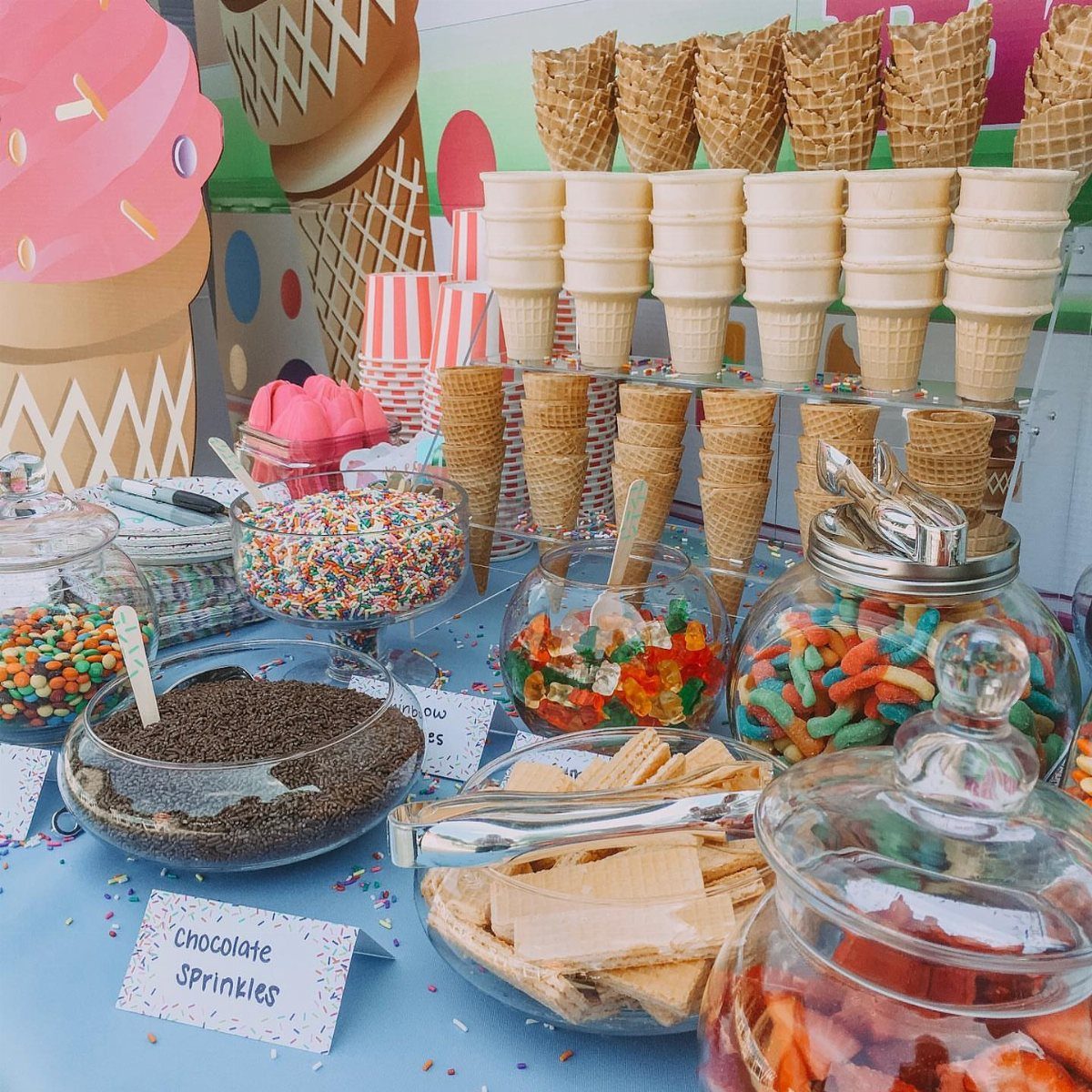Ice cream bar with waffle cones, sprinkles, and gummy candies on blue tablecloth.