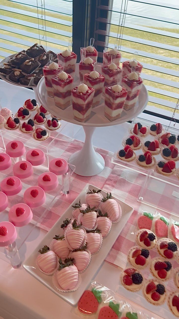 Dessert table with pink mousse cakes, strawberry cakes, and chocolate-dipped strawberries on checkered cloth.