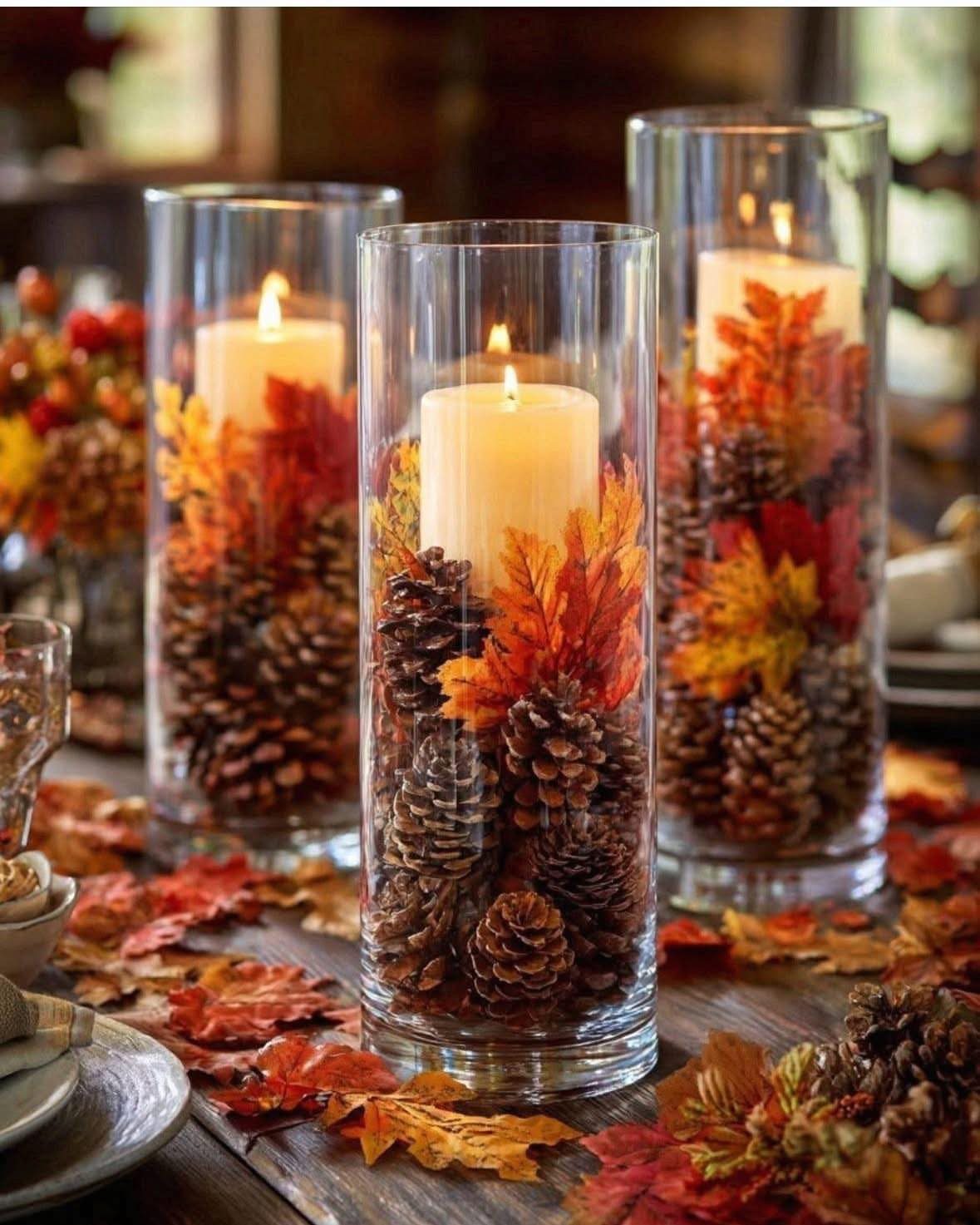 Lit white pillar candles in glass vases with pinecones and autumn leaves on table