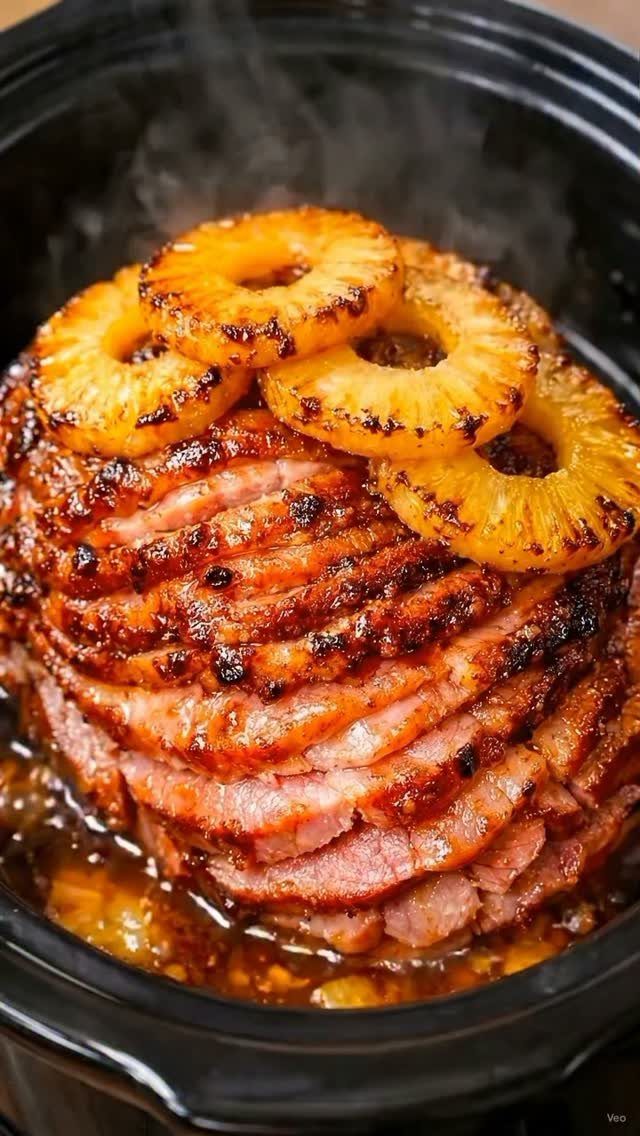 Glazed spiral-sliced ham topped with pineapple rings inside a black slow cooker pot.
