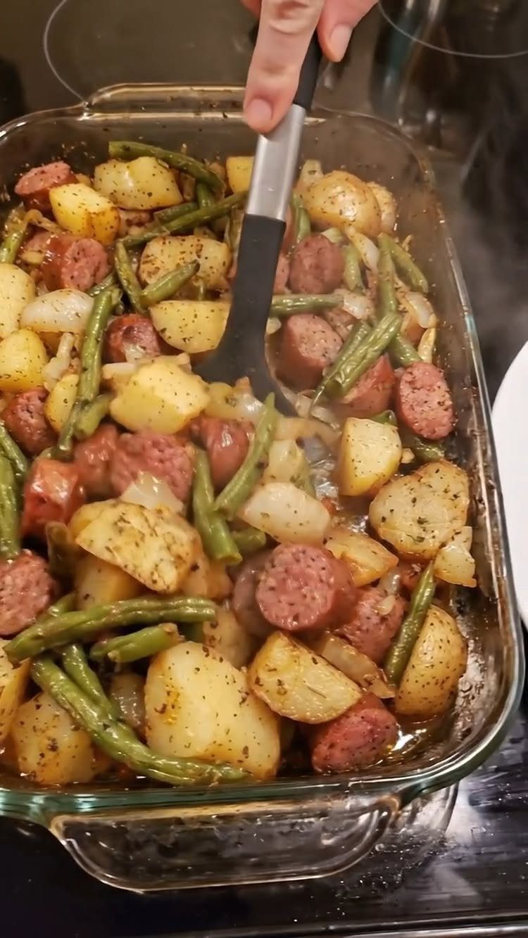 Sausage, potato, and green bean casserole in a glass baking dish being stirred.
