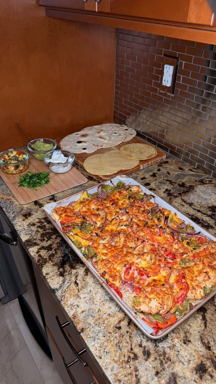 Sheet pan chicken fajitas with melted cheese, tortillas, and side dips on granite.