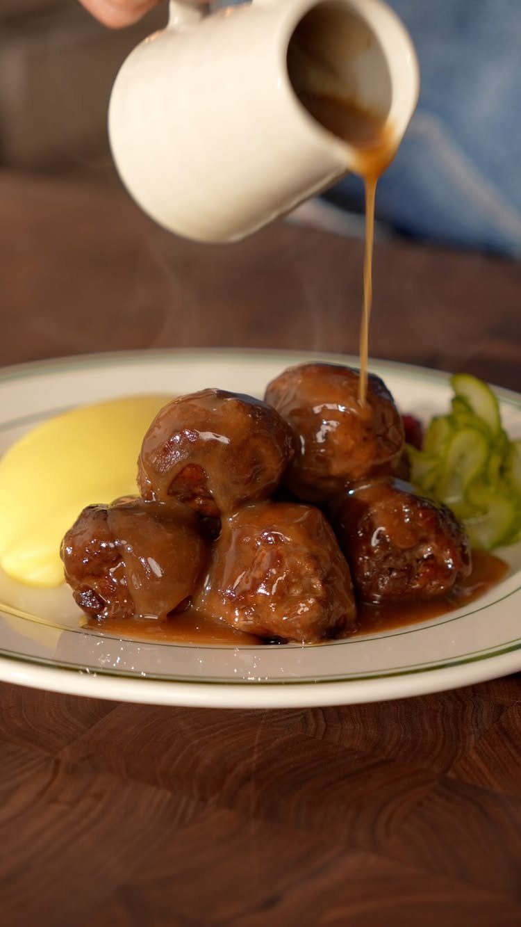 Brown gravy poured over glazed meatballs with mashed potatoes and cucumbers on a plate.