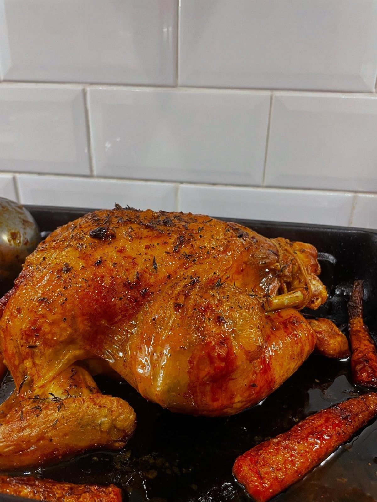 Whole roasted chicken with carrots in a black pan against white tiles
