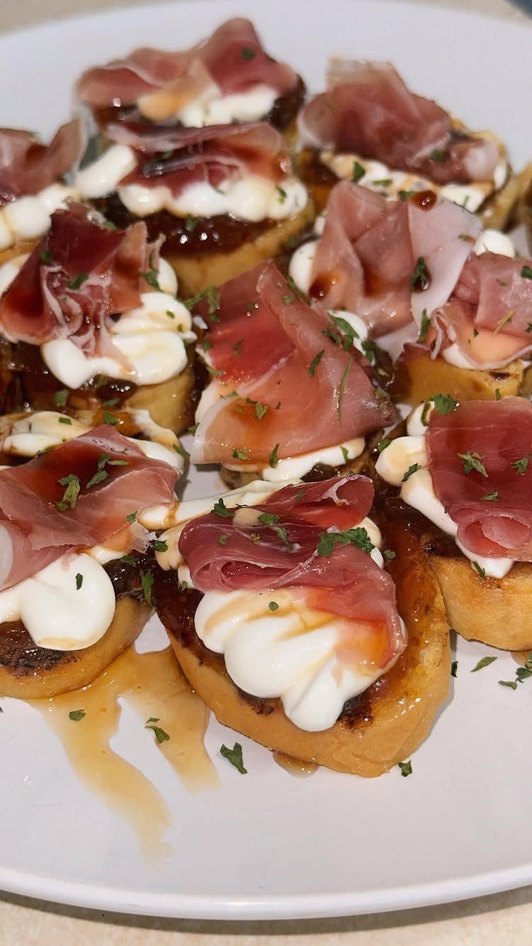 Toasted baguette slices topped with creamy white cheese and prosciutto on a white plate.