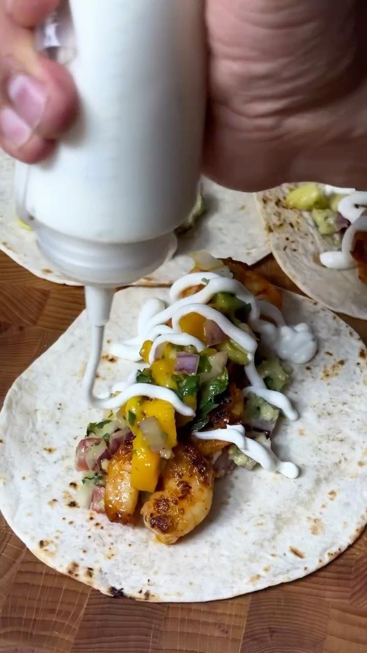 Grilled shrimp tacos with mango salsa being drizzled with white cream sauce.