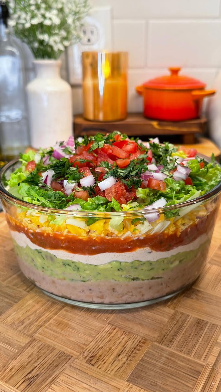 Seven-layer dip in a glass bowl topped with fresh vegetables on wooden board.