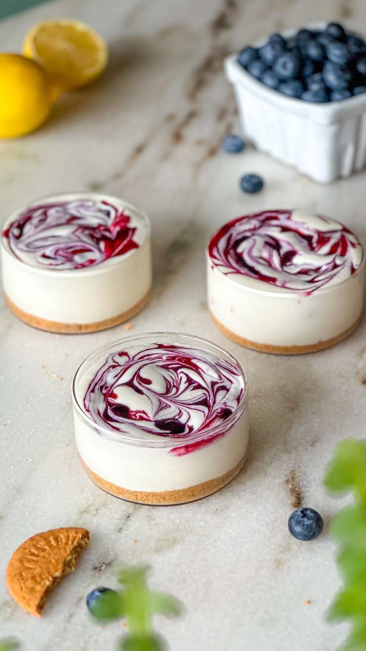 Three mini cheesecakes with berry swirls in glasses on a marble countertop.