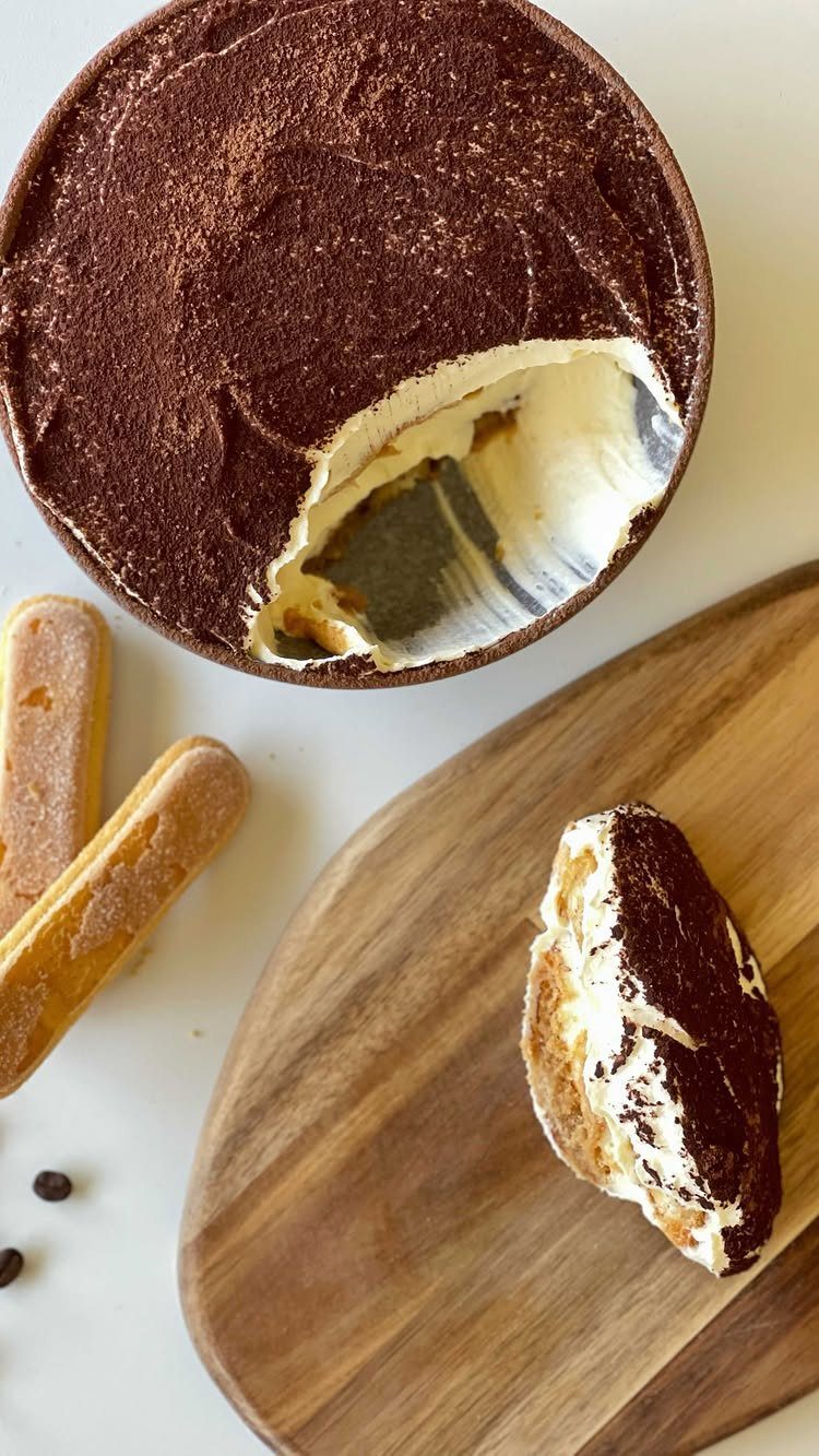 Tiramisu cake with cocoa powder, a slice on a wooden board and ladyfingers.