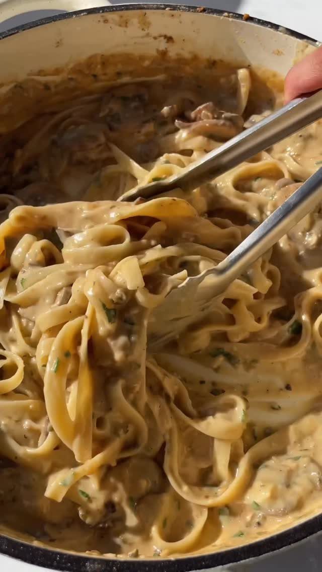 Creamy mushroom pasta with wide noodles being lifted by metal tongs in a pot.