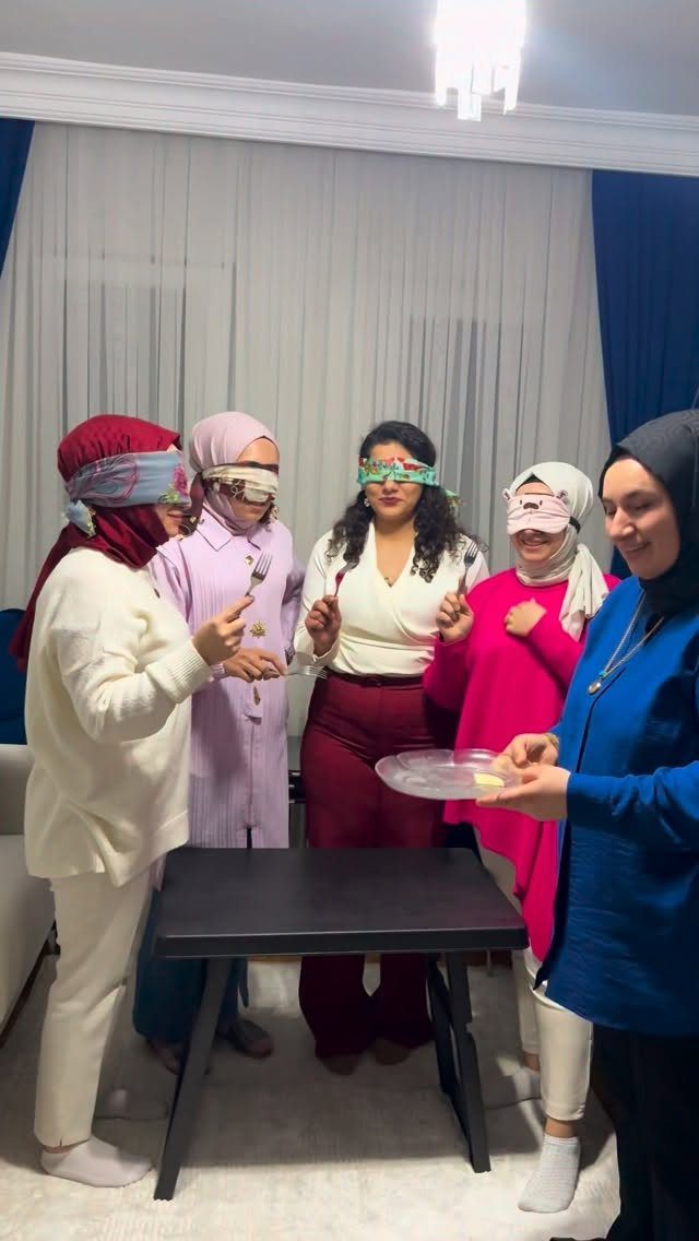 Blindfolded women holding forks while another woman holds a plate of food.