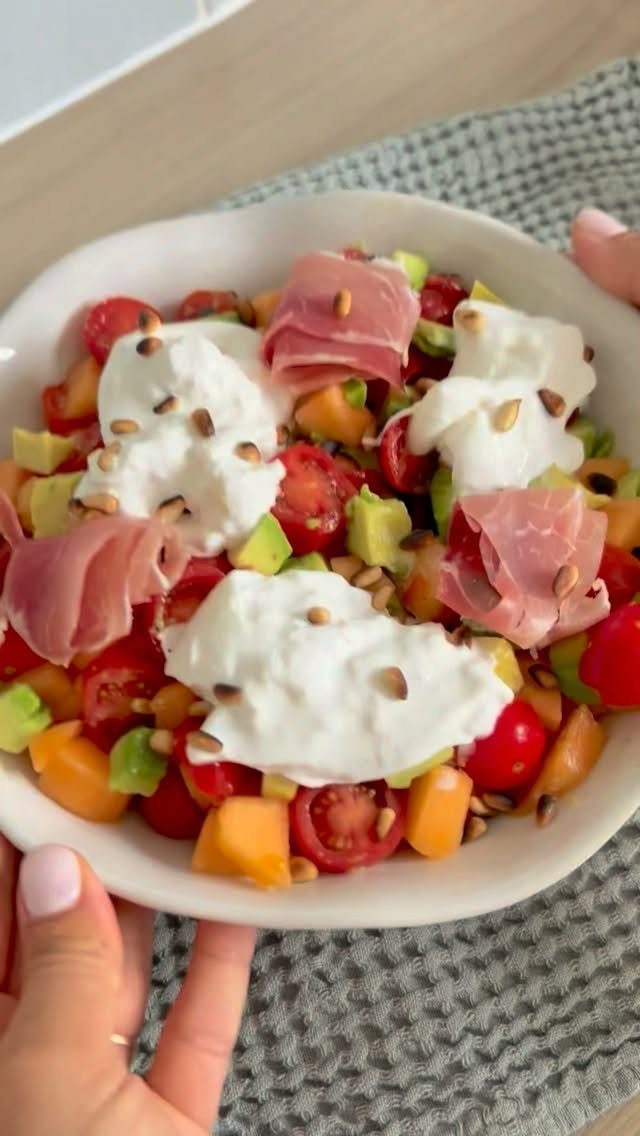 Melon salad with cherry tomatoes, avocado, prosciutto, and creamy white cheese in bowl.