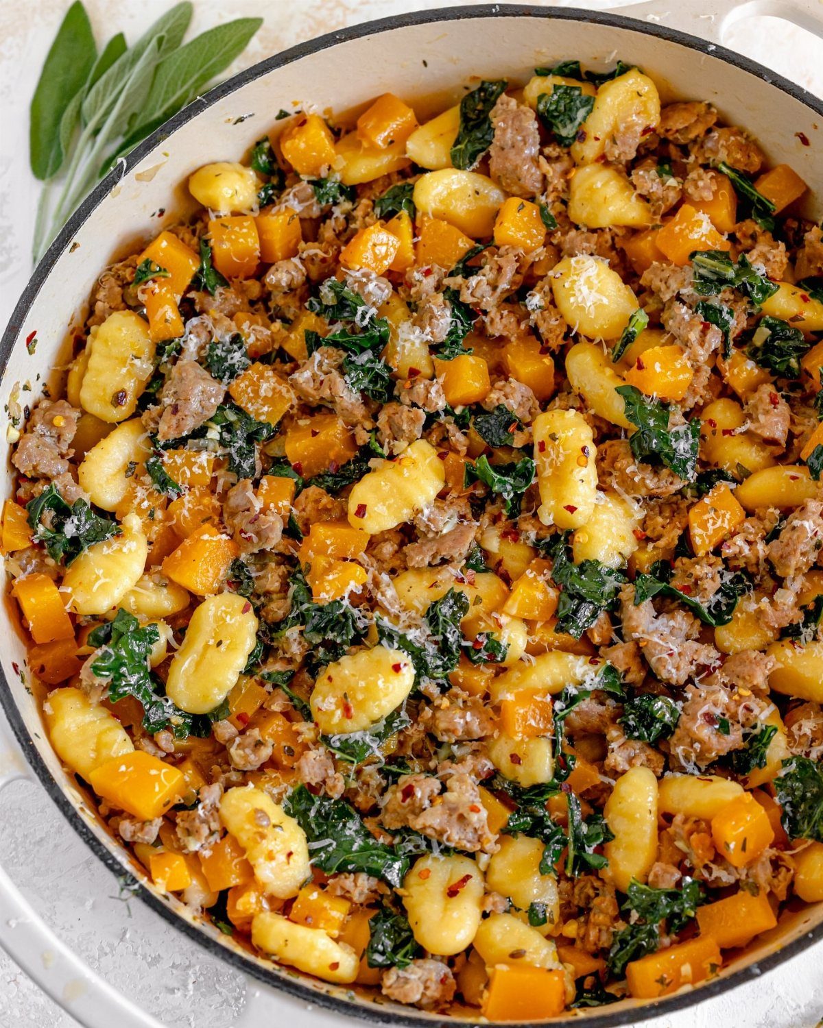 Gnocchi with squash, sausage, and kale in a white pot with sage leaves.