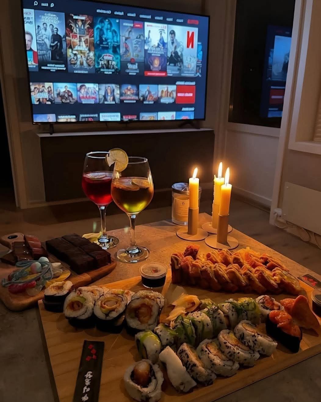 Sushi platter with cocktails and candles on a table before a television screen.