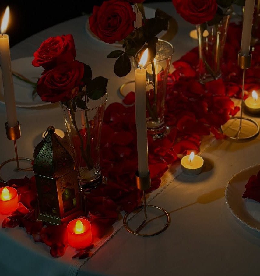 Romantic dinner table arrangement with red roses, petals, and glowing candles on white cloth