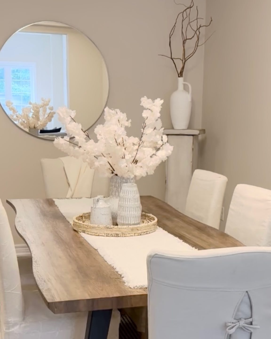 White textured vases with blossoms on a woven tray atop a wooden table.