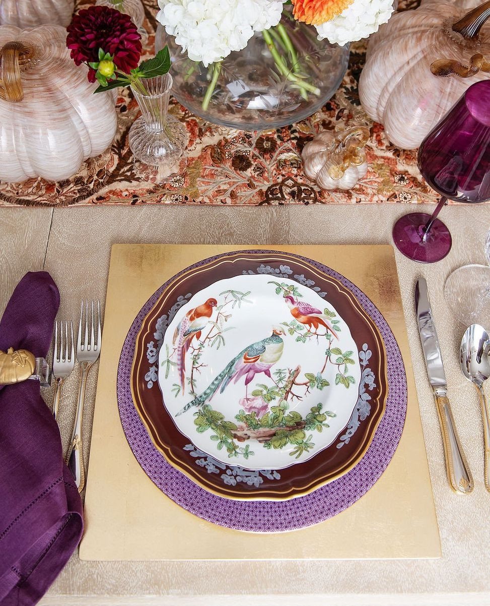 Layered dinnerware with bird illustrations on a gold placemat with purple accents.
