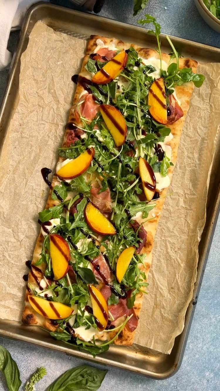 Rectangular flatbread pizza topped with prosciutto, peach slices, arugula, and balsamic glaze.