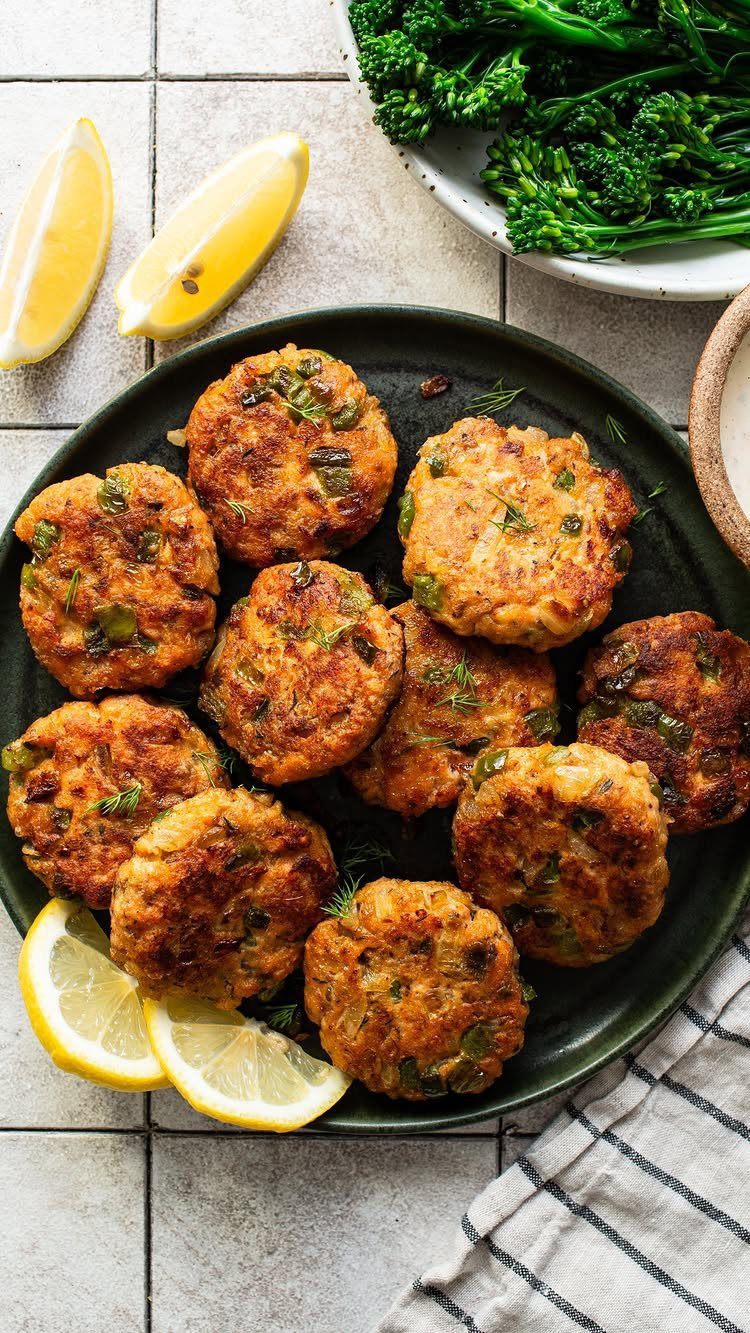 Golden-brown salmon patties on a green plate with lemon wedges and steamed broccolini.