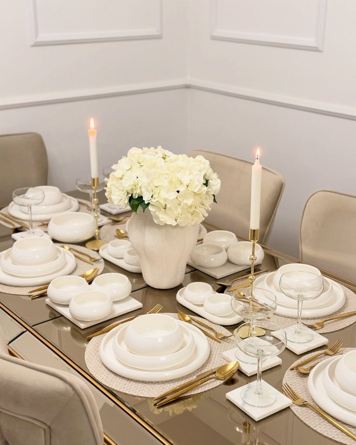 White plates and gold cutlery on a glass table with white flowers.