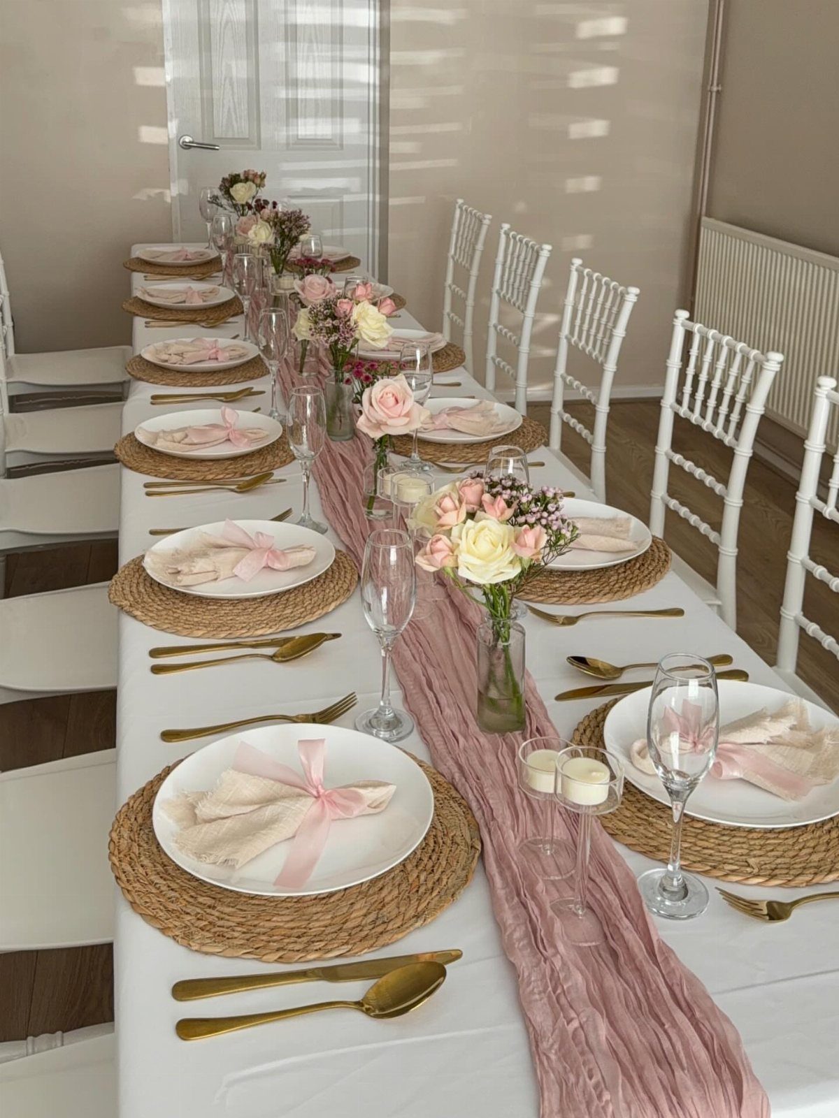 Long white table with beige woven placemats, pink runner, and floral centerpieces.