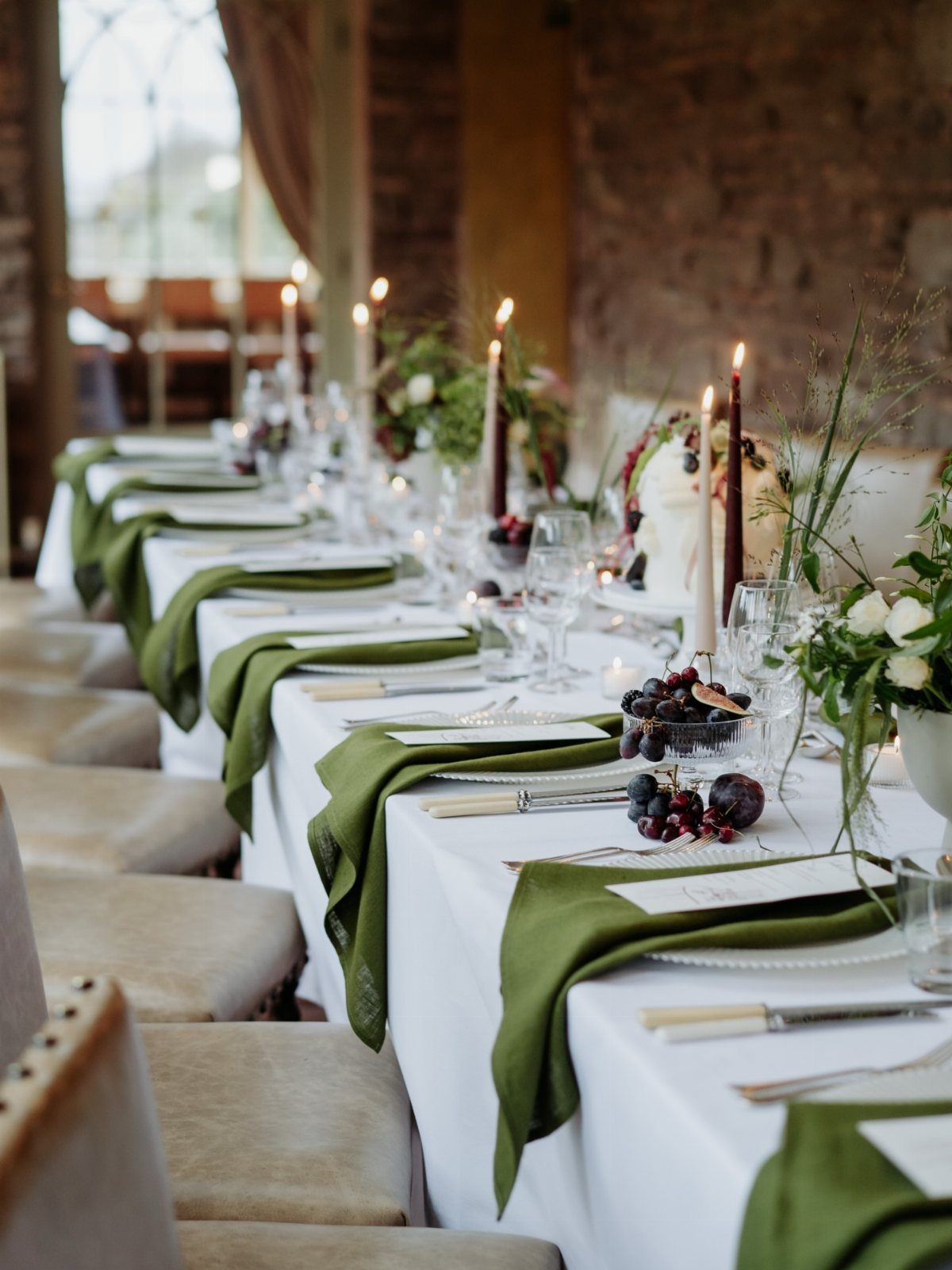 Long table setting with white cloth, green napkins, burgundy candles, and fresh greenery.