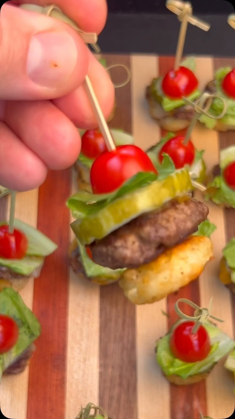 Mini sliders with cherry tomatoes and pickles on a striped wooden serving board.