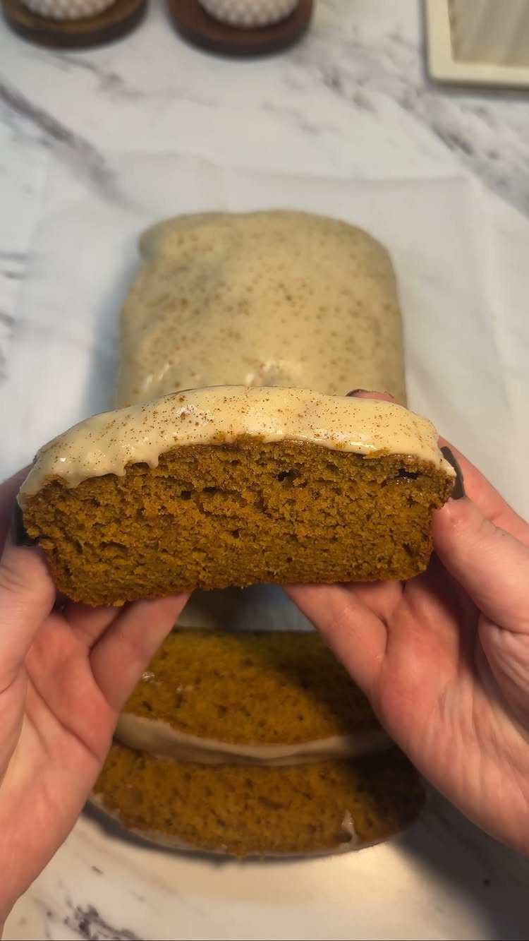 Slice of glazed pumpkin bread held by hands with a loaf behind it.