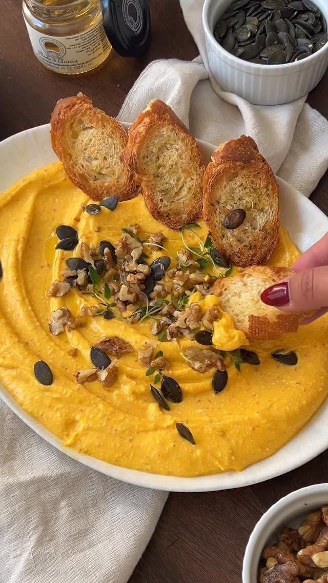 Orange pumpkin hummus in a white bowl topped with seeds and toasted baguette.