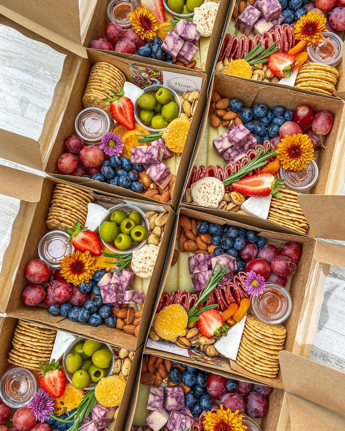 Charcuterie boxes filled with crackers, various cheeses, fresh fruits, olives, and sliced meats.