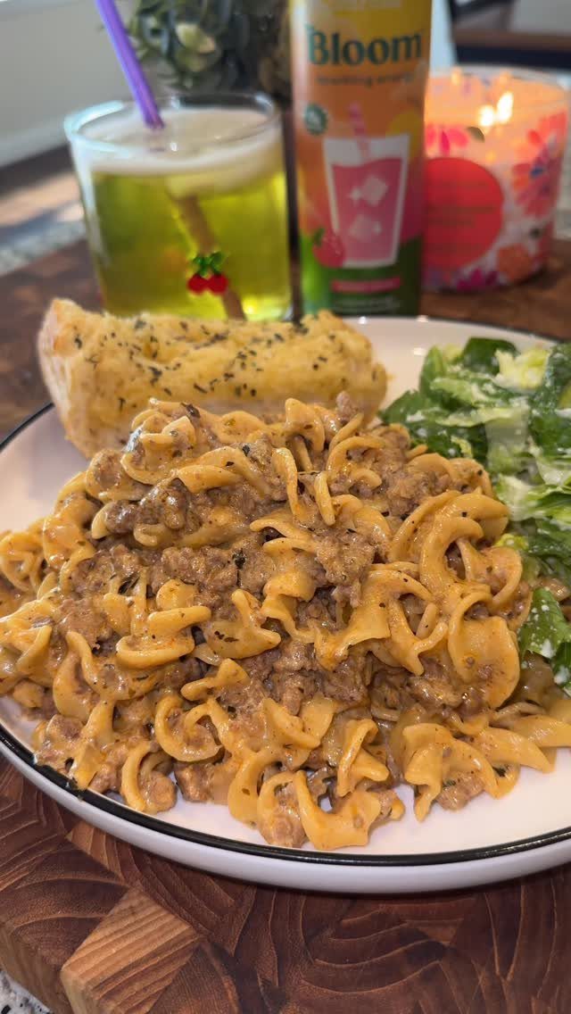 Beef stroganoff with egg noodles, garlic bread, and salad on a white plate.