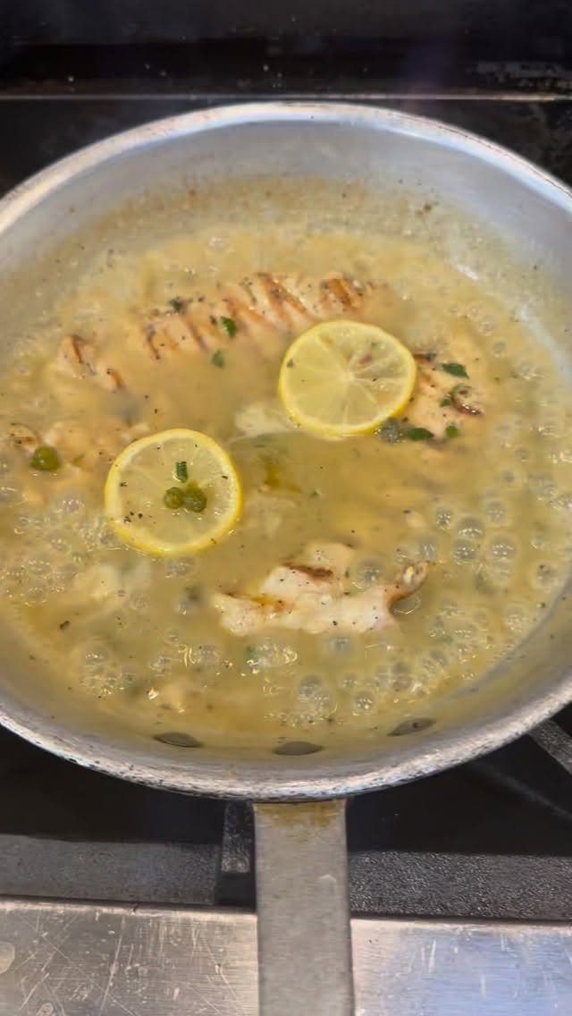 Chicken breasts simmering in a lemon caper sauce in a silver skillet.